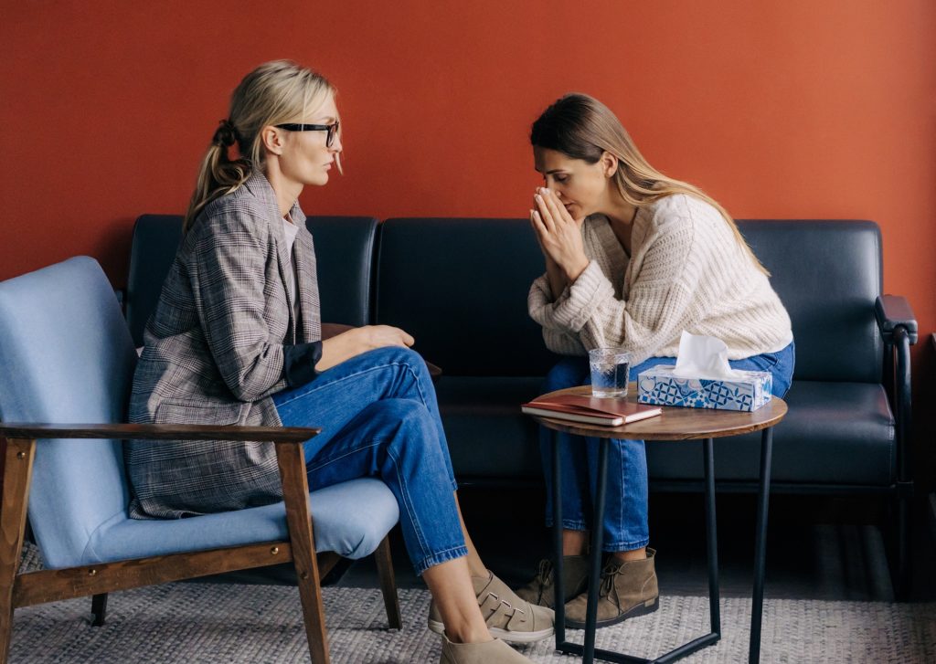 Crying depressed female client sitting on couch in therapist's workshop talking about trauma.